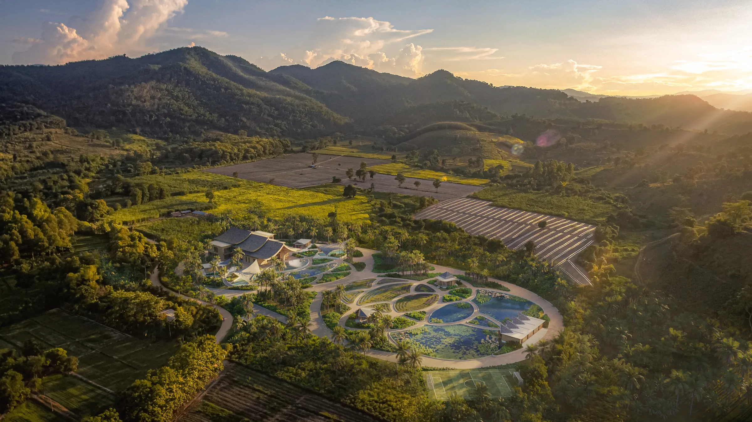 Aerial 3D rendering of a retreat community masterplan in a green Thai valley, a low-density campus of pavilions, curving paths, gardens and ponds ringed by forested mountains, with a low sun on the horizon.