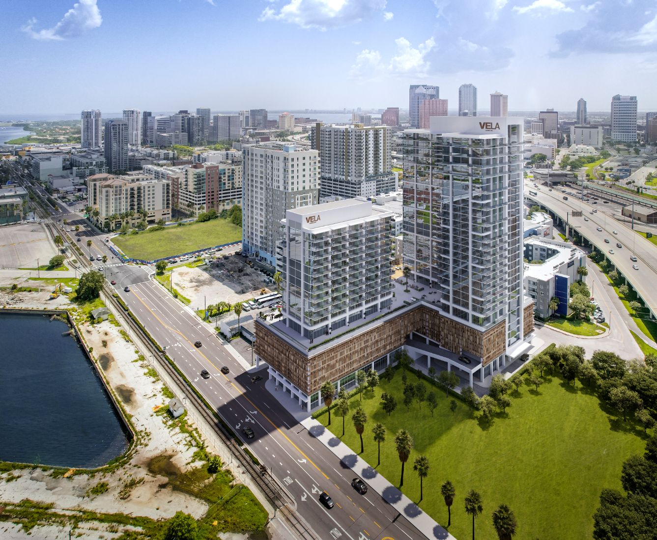 Aerial 3D rendering of the VeLa twin mixed-use towers in Tampa, rising from a podium on a waterfront site beside a highway and palm-lined boulevard.