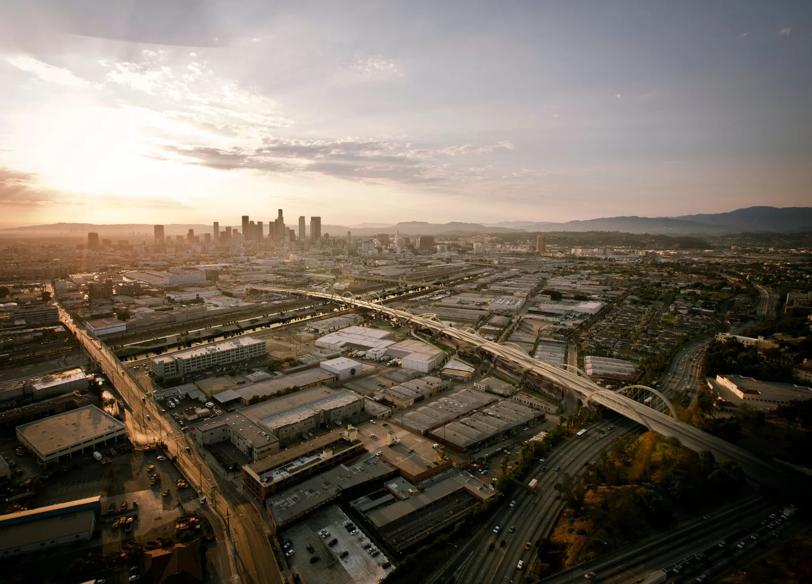 Aerial 3D rendering looking across the industrial flats of Los Angeles at golden hour, with the Sixth Street Viaduct curving toward the Downtown skyline on the horizon.